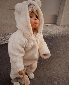 a toddler in a white teddy bear costume standing on the sidewalk with her hands behind her head