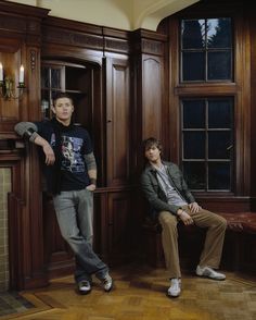 two young men sitting on wooden chairs in front of large wood paneled walls and windows