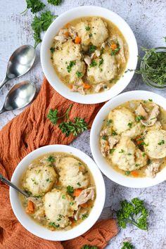 three white bowls filled with chicken and dumplings next to silver spoons on an orange napkin