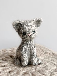 a small gray knitted kitten sitting on top of a white blanket and looking at the camera