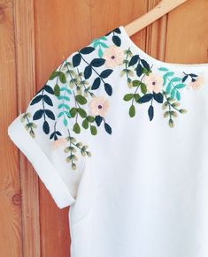 a white shirt with flowers and leaves on it hanging from a hanger against a wooden wall