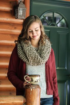 a woman standing next to a log holding a coffee mug in her right hand and wearing a scarf around her neck