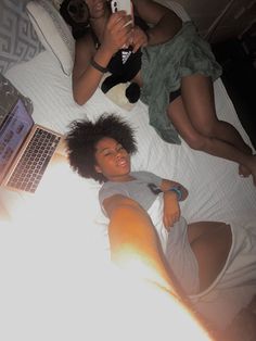 two women laying on a bed looking at a cell phone and laptop computer, with one holding a stuffed animal