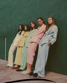 four women in pastel outfits leaning against a green wall with their hands on their hips