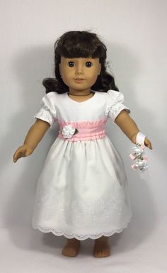 the doll is wearing a white dress with pink trimmings and a flower in her hand