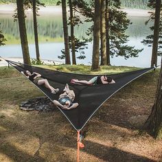 two people laying in a hammock on the ground near some trees and water