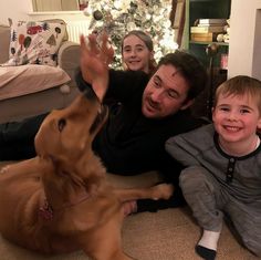 a man, woman and child sitting on the floor with a dog in front of a christmas tree