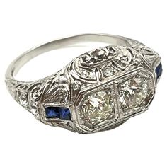 This vintage, Edwardian ring is handmade and set in platinum as is stamped inside the shank. Elegant, milgrain detailing and scroll work make this piece stand out! It features two Old European-cut diamonds as its center stones and a variation of rose and full-cut accent diamonds along the shoulders and body of the ring. The full-cut diamonds are most likely new additions to the piece. Two sapphires adorn each shoulder for a total of four colored stones. The total diamond weight is approximately Antique Platinum Three Stone Diamond Ring, Antique Platinum Three-stone Jewelry, Art Deco Three Stone Platinum Jewelry, Antique Platinum Three Stone Rings, Antique Three Stone Platinum Rings, Heirloom Three Stone Platinum Ring, Heirloom Platinum Three Stone Ring, Heirloom Platinum Three-stone Ring, Heirloom Three-stone Platinum Ring