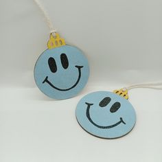 two blue ornaments with smiley faces hanging from string on white surface, one has a gold crown on it and the other has a smiling face