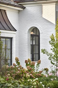 a white brick house with lots of flowers in the foreground and bushes to the side