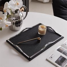 a white table topped with a black tray and a silver vase filled with flowers on top of it