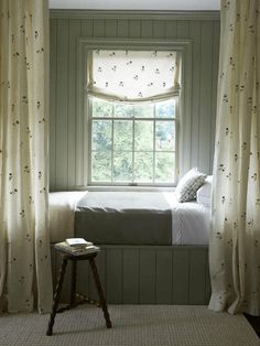 a bed sitting under a window next to a wooden stool in front of a window