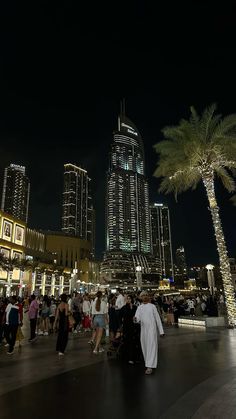 many people are walking around in the city at night with tall buildings and palm trees