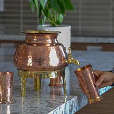 a person is pouring water into a copper pot on a marble table with two cups