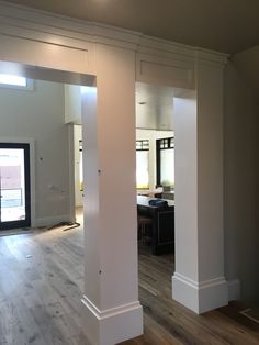 an empty room with white columns and hard wood flooring in the center, looking into the kitchen