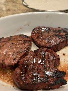 three steaks are in a white bowl on a counter top next to some sauce