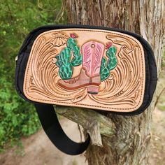 This bag has a hand-tooled leather patch featuring western floral carving and a cowgirl boot surrounded by cacti. It can be worn crossbody or around the waist. There is a large zippered opening and a small zippered pocket on the back. Western Hand Tooled Travel Bag, Western Style Hand-tooled Travel Bag, Western Leather Bags With Concho Details, Western Style Hand Tooled Shoulder Bag For Travel, Western Style Crossbody Bags For Western-themed Events, Western Style Travel Bag With Leather Backing, Western Style Crossbody Shoulder Bag For Travel, Tooled Leather Belts, Cowgirl Boot