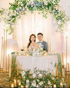 a man and woman sitting at a table in front of a floral arch with candles