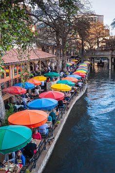 many people are sitting under umbrellas on the side of a river while others sit at tables