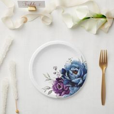 a white plate with blue flowers on it next to gold utensils and napkins