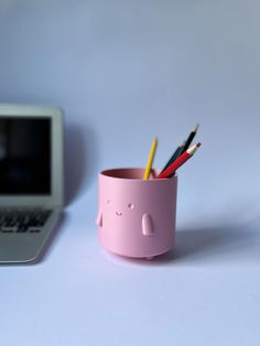 a pink cup with pencils in it next to a laptop on a white table