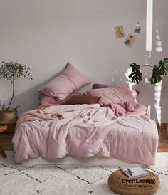 an unmade bed with pink linens and pillows in a white room next to potted plants