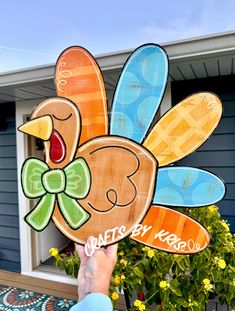 a person holding up a wooden turkey sign in front of a blue house with yellow flowers