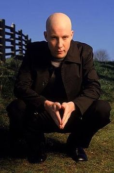 a bald man sitting on top of a grass covered hill next to a wooden fence