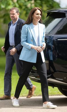 a woman in a blue blazer and black pants walking next to a car with the door open