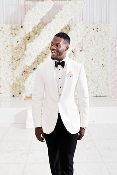 a man in a white tuxedo smiles at the camera while standing next to a flower wall