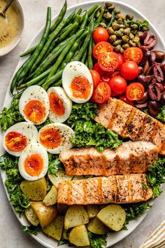 a white plate topped with meat, vegetables and hard boiled eggs next to olives