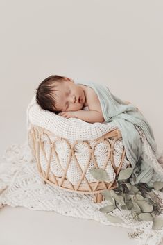 a newborn baby is sleeping in a basket on a white blanket with greenery around it