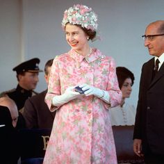 an older woman in a pink dress and white gloves smiles as she stands next to another man