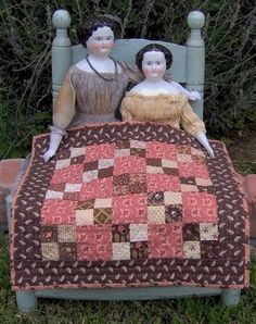 two dolls sitting on top of a quilted bed in the middle of some grass