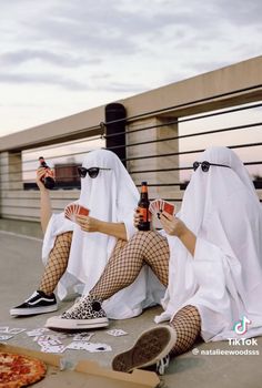 two women dressed in white are sitting on the ground with beer and playing cards together