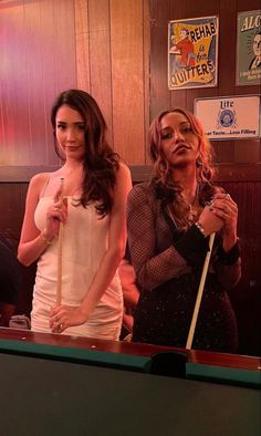 two women standing next to each other in front of a pool table with cues