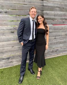 a man and woman standing next to each other in front of a wooden fence with grass