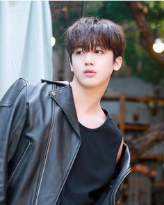 a young man in a black shirt and leather jacket standing next to a tree with lights hanging from it