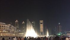 people are standing around in front of a fountain at night with buildings and skyscrapers in the background