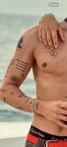 a shirtless man with tattoos on his arm and chest standing next to the ocean