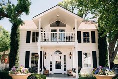 a white house with black shutters on the front and side windows, surrounded by greenery