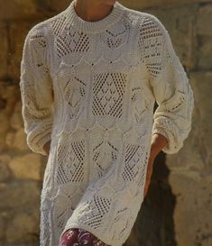 a woman wearing a white sweater and patterned pants is standing in front of a stone wall