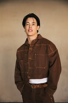 a young man standing in front of a wall wearing a brown jacket and white shirt