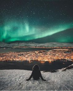 a person sitting in the snow looking at an auroral display over a small town