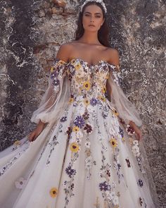 a woman in a white dress with flowers on it standing next to a stone wall