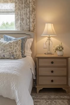 a white bed sitting under a window next to a night stand and lamp on top of a dresser