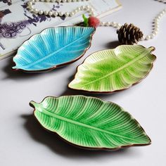 three leaf shaped dishes sitting on top of a white table next to a pine cone