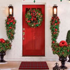 two christmas wreaths on top of urns in front of a red door with lights