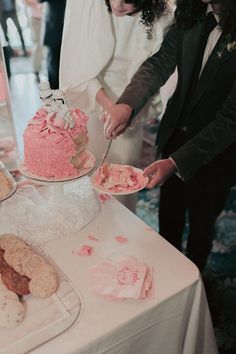 two people standing at a table with cakes and cookies on it, one is cutting the cake