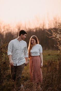 a young man and woman walking through tall grass in an open field at sunset with the sun setting behind them